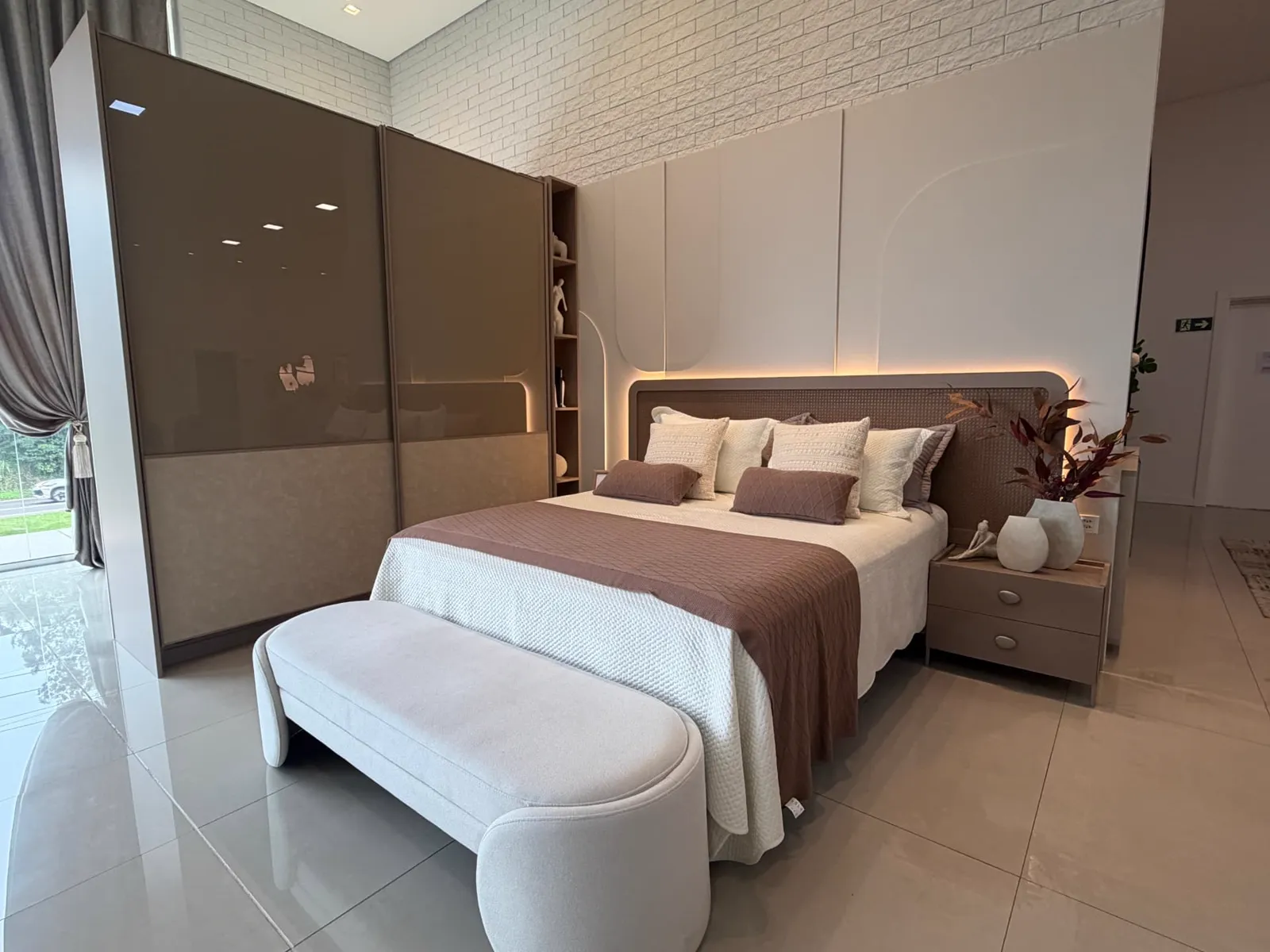 Suíte de casal do showroom com guarda-roupa de vidro reflecta, cabeceira em palhinha e iluminação embutida